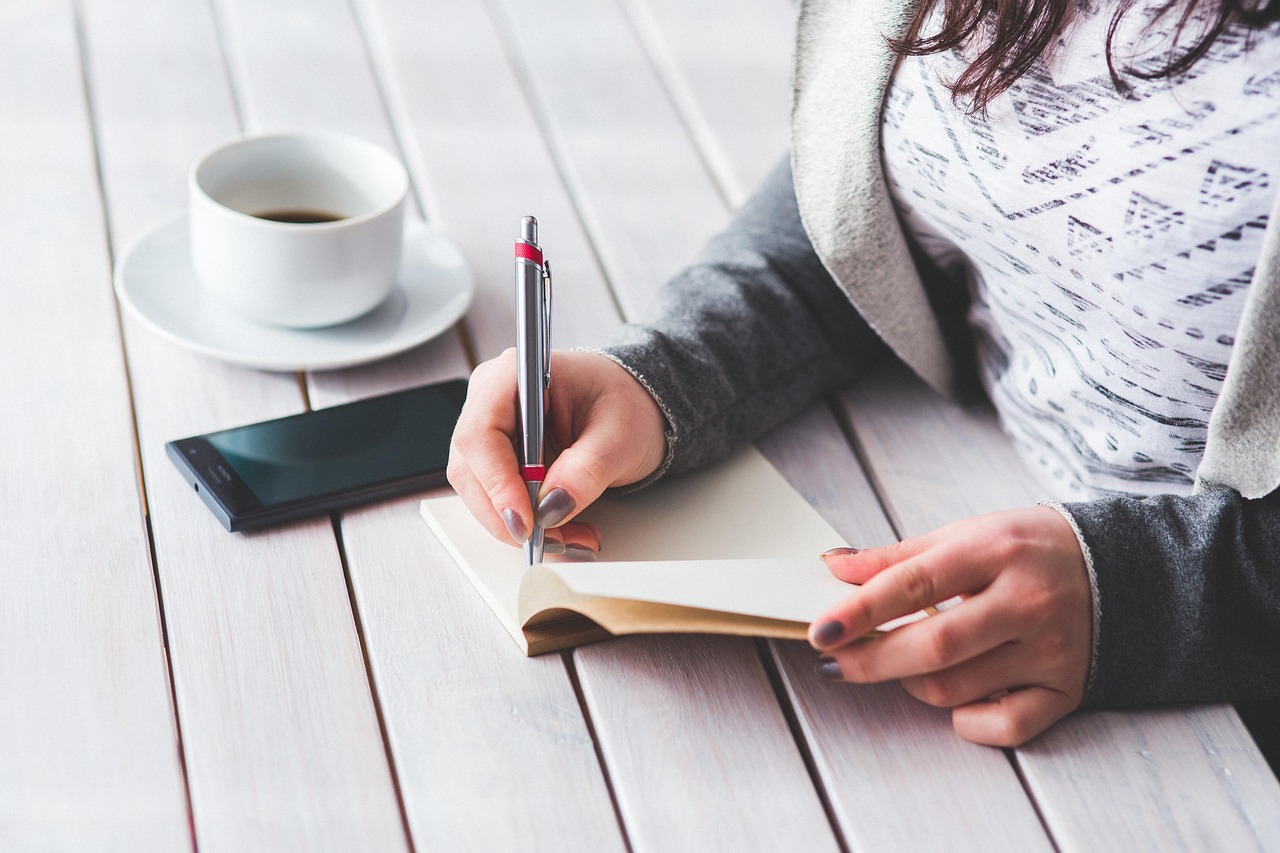 Vrouw schrijft notities in een schrift aan een tafel met een kop koffie en een smartphone.