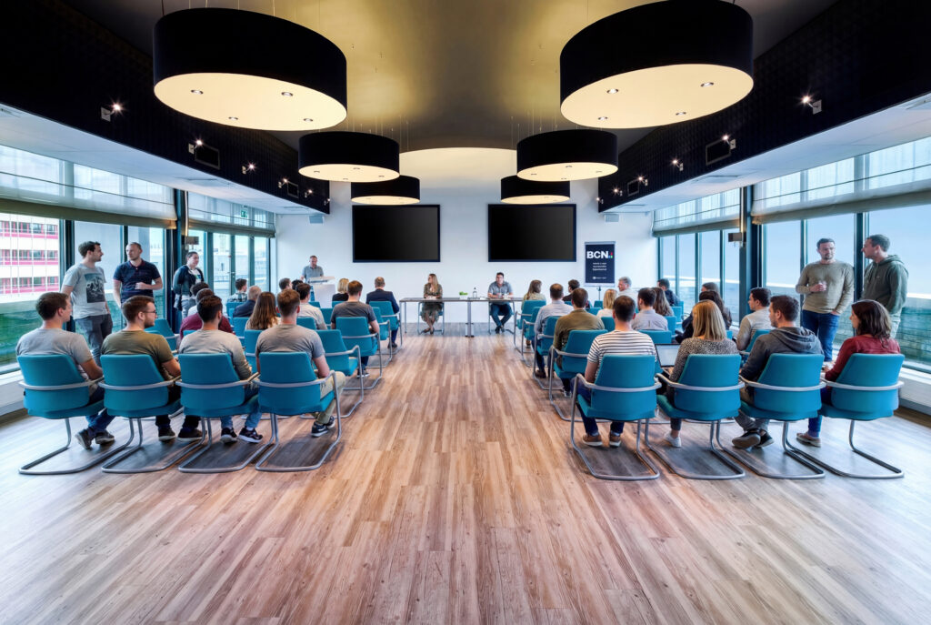 Groep mensen volgt een training in een moderne, ruime zaal met grote ramen en blauwe stoelen.
