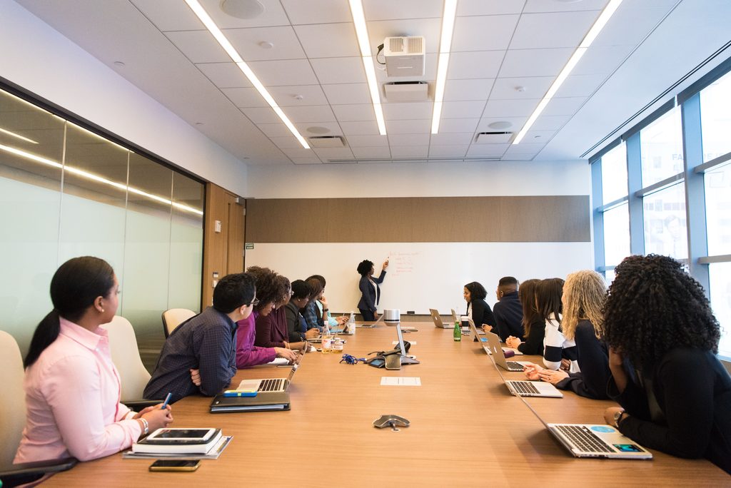 Mensen tijdens vergadering acher laptop kijkend naar mevrouw die voor het whiteboard staat.